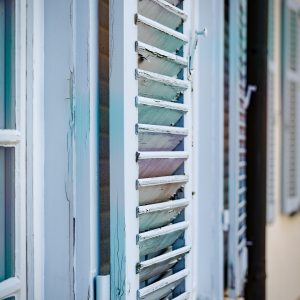 Nahaufnahme eines alten, halb geöffneten Fensterladens aus Holz mit abblätternder Farbe in Pastelltönen. Im Hintergrund sind weitere Fenster und Läden entlang einer Hausfassade unscharf zu sehen.