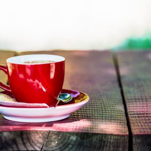Eine rote Kaffeetasse auf Untertasse mit Löffel und Zuckerstückchen steht auf einem rustikalen Holztisch; der Hintergrund ist hell und unscharf, mit grünlichen Farbflecken und Sonnenlicht.